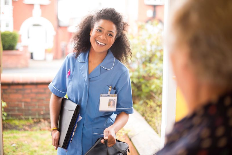 A smiling nurse-trained carer arriving at a client's home with a folder of notes.