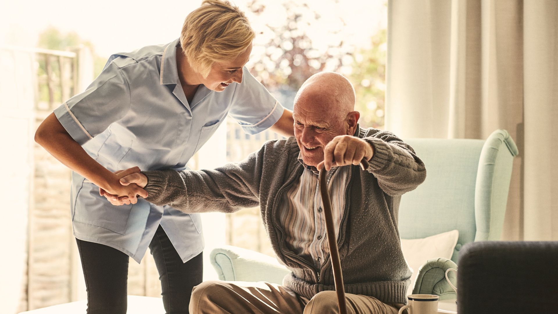 A nurse-trained companion supporting an older gentleman to stand from a sofa in his own living room.